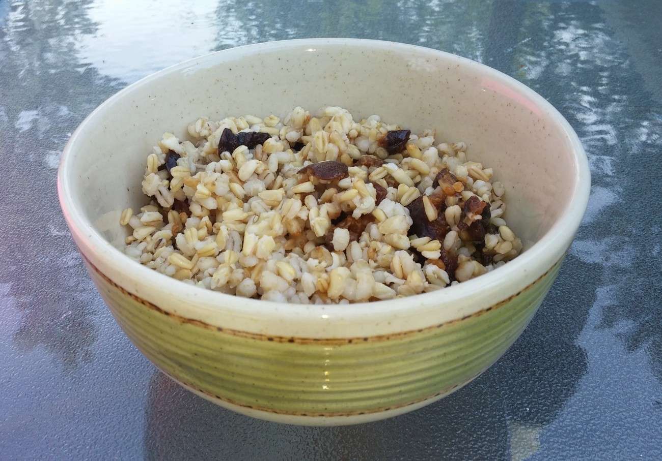 A ceramic bowl with a green exterior, filled with a delightful, plant-based mixture of grains and dark bits, likely dried fruit or nuts, rests on a glass surface. Curated by a dietitians expertise, the grains are lightly cooked and appear fluffy, embracing vegetarian goodness.
