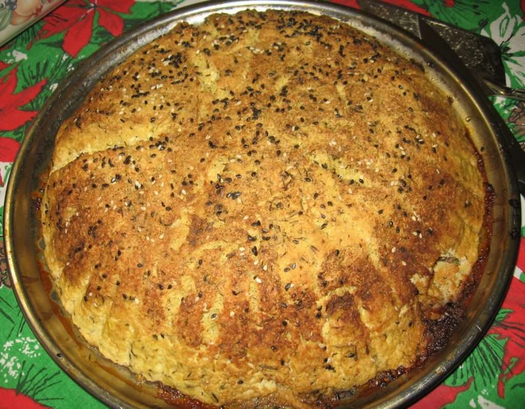 A golden-brown, round bread rests on a metal baking tray, topped with sesame seeds and herbs. This plant-based delight has a dense, crusty texture and sits elegantly on a red and green tablecloth.