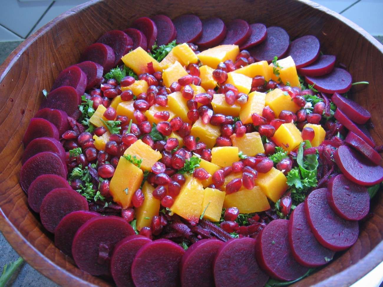 A wooden bowl cradles a vibrant, plant-based salad. Sliced beets form a border around the edge, while orange mango chunks, red pomegranate seeds, and green parsley are artfully arranged on a bed of spinach leaves.