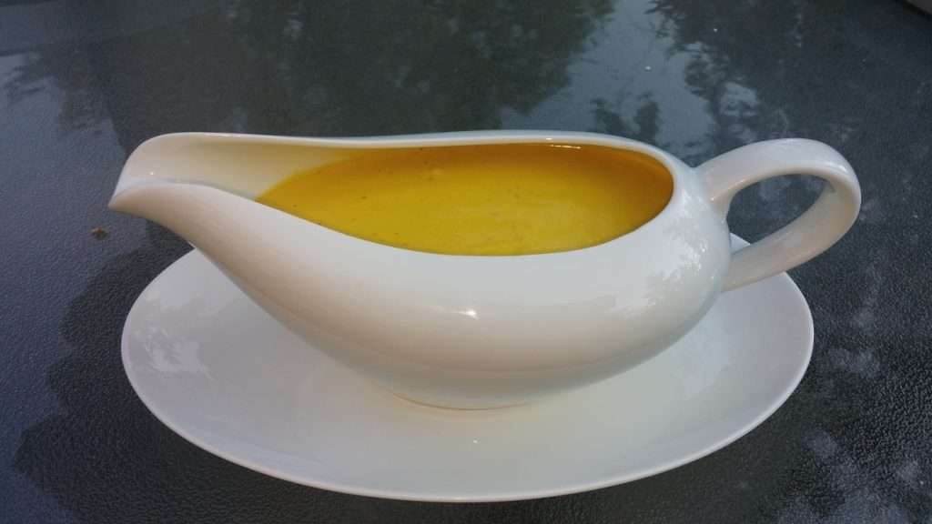 A white gravy boat filled with plant-based yellow sauce or dressing sits on a matching saucer. The surface around it appears to be a glass table, reflecting some of its surroundings.