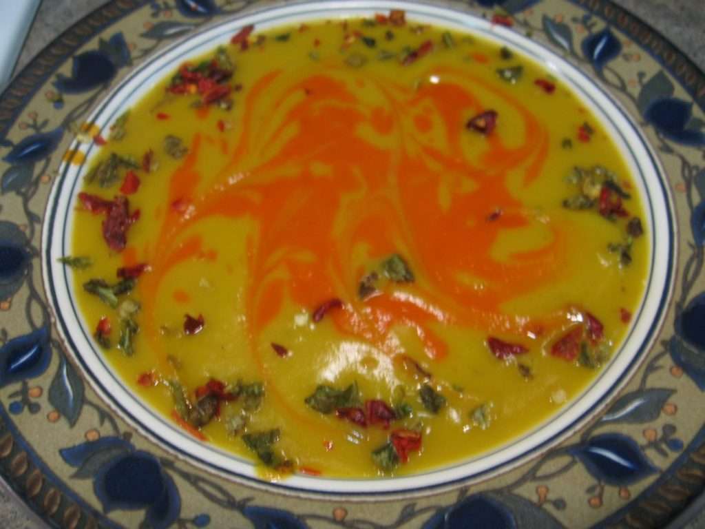 A decorative plate filled with vegan yellow lentil soup, topped with a swirl of red chili oil and garnished with chopped herbs and spices. The soup is set on a floral-patterned border, offering a feast for the eyes and palate alike.