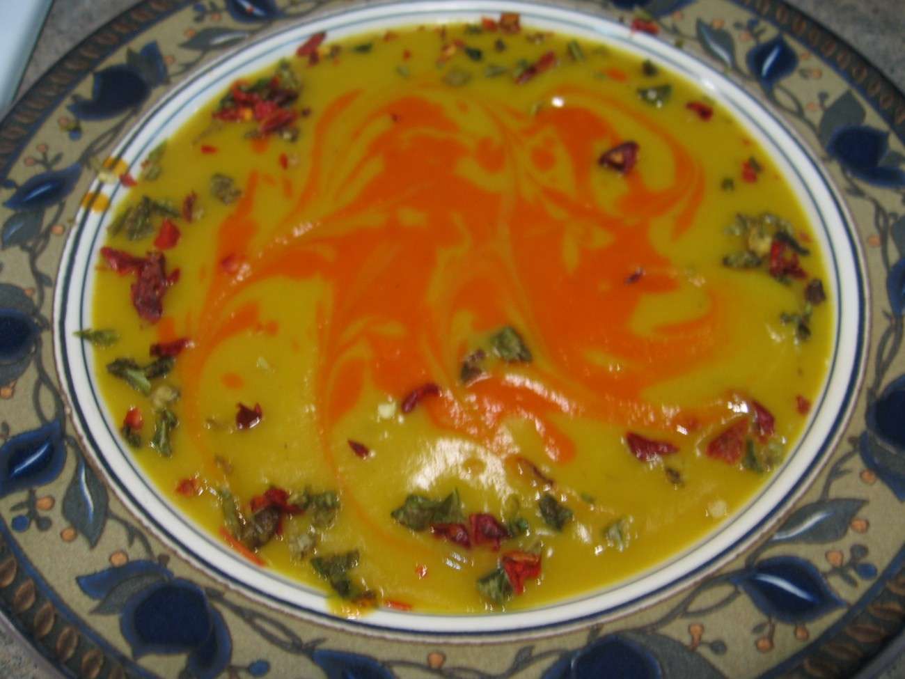 A decorative plate filled with vegan yellow lentil soup, topped with a swirl of red chili oil and garnished with chopped herbs and spices. The soup is set on a floral-patterned border, offering a feast for the eyes and palate alike.