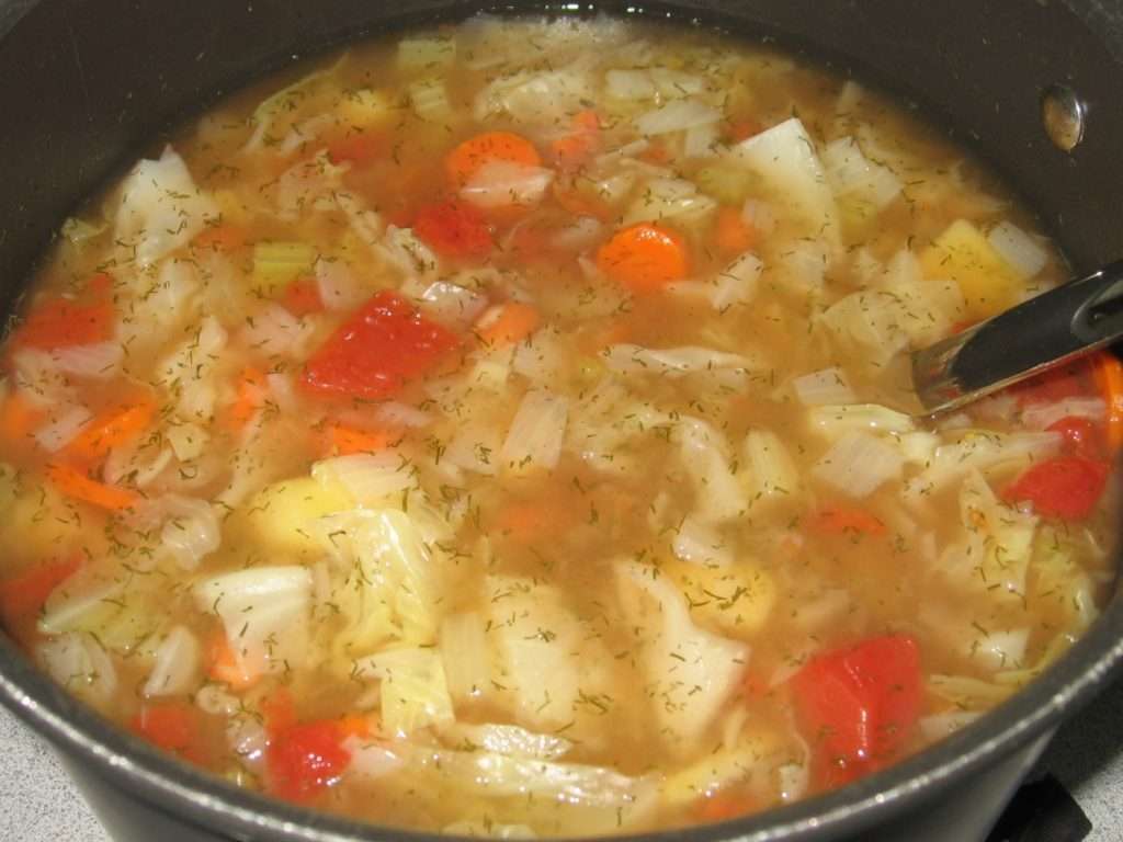 A pot filled with plant-based cabbage soup, featuring chopped cabbage, carrots, red bell peppers, and potatoes in a clear broth. A ladle is partially submerged in this vegan delight. The surface is sprinkled with herbs.