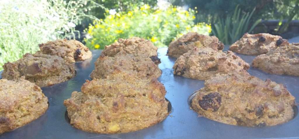 Freshly baked vegan muffins with chunky tops in a muffin tray outdoors. The background features a garden with lush green foliage and vibrant yellow flowers, creating a sunny and natural setting.