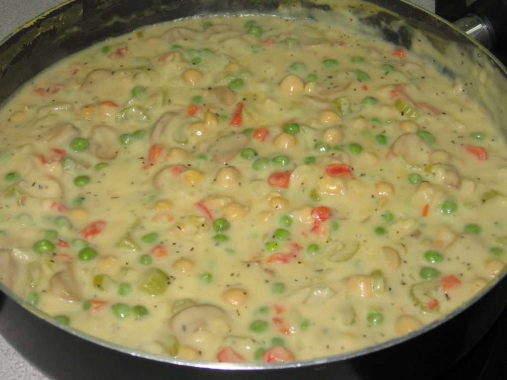 A pot filled with a creamy, plant-based vegetable stew, featuring diced carrots, peas, chickpeas, mushrooms, and celery in a thick sauce. This vegan delight appears well-seasoned with visible herbs and spices.
