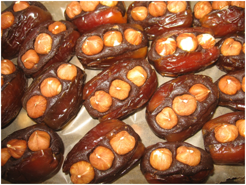 Close-up of vegan dates stuffed with hazelnuts, arranged on a tray. The dark brown, glossy dates are split open to reveal two to three hazelnuts inside, offering a delightful contrast in texture and color.