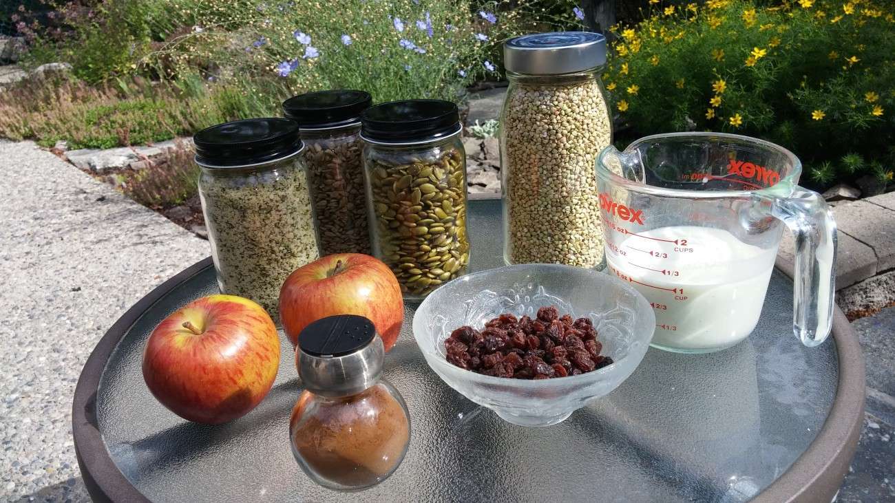 A glass table outdoors showcases plant-based ingredients for cooking: a bowl of raisins, two apples, a jar of cinnamon, three jars with seeds or grains, a jar of buckwheat, and a measuring cup with milk. Blossoming flowers and lush plants add to the picturesque vegetarian vibe in the background.
