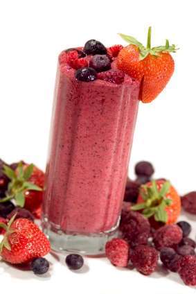 A tall glass of vibrant pink mixed berry smoothie, perfect for a vegetarian delight, is garnished with whole strawberries and surrounded by scattered strawberries, raspberries, and blueberries on a white background. Enjoy the visible berry bits in this plant-based treat.