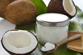A jar of plant-based coconut oil sits on a large green leaf, surrounded by whole and halved coconuts. A wooden spoon with a small amount of this vegan oil rests on the leaf, highlighting the natural setting.
