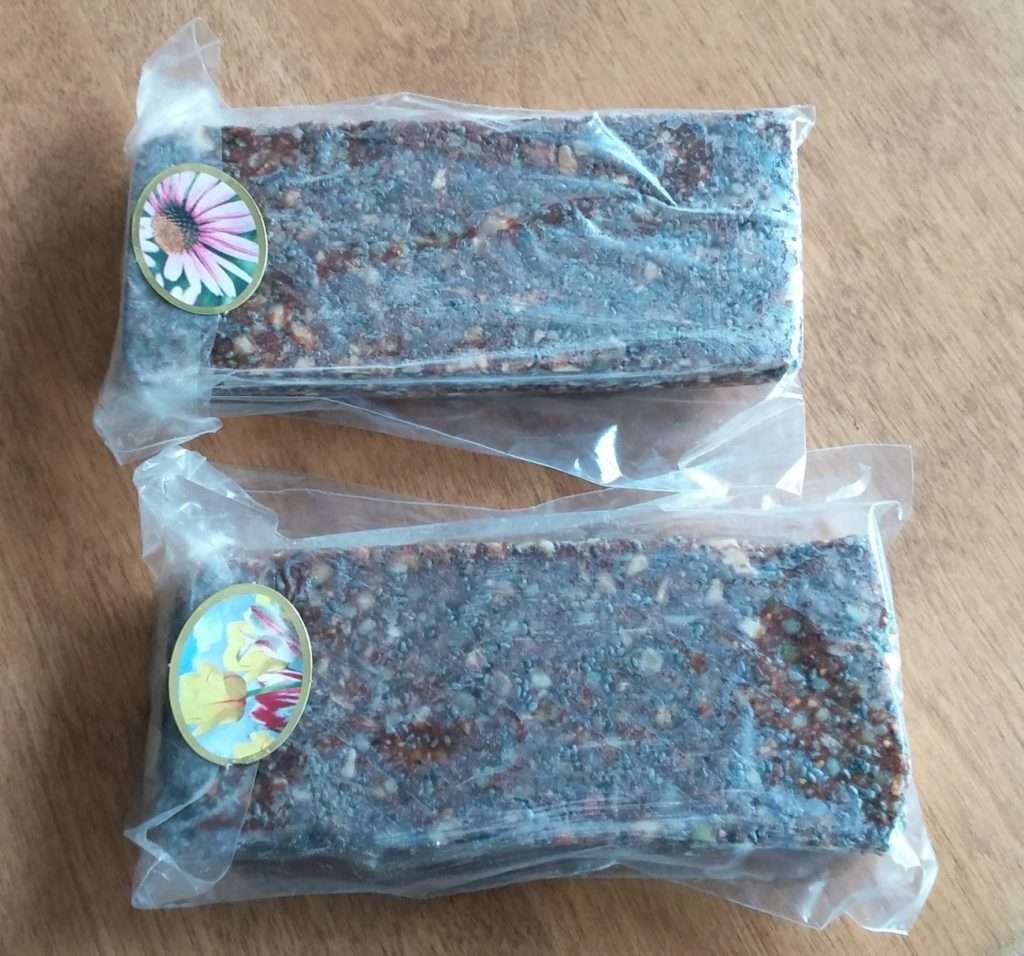 Two rectangular, vacuum-sealed packets of brown speckled food rest on a wooden surface. Each vegetarian packet features a circular sticker adorned with floral images, one showcasing a pink flower and the other yellow flowers, highlighting its plant-based nature.