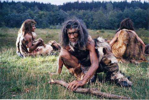 A group of three people dressed as prehistoric humans sits on grass in a woodland area. Embracing a natural lifestyle, they don animal skins and have unkempt hair. One person crouches with a stick, while the others sit back, reminiscent of our ancestors simpler times before dietitians and vegan trends emerged.