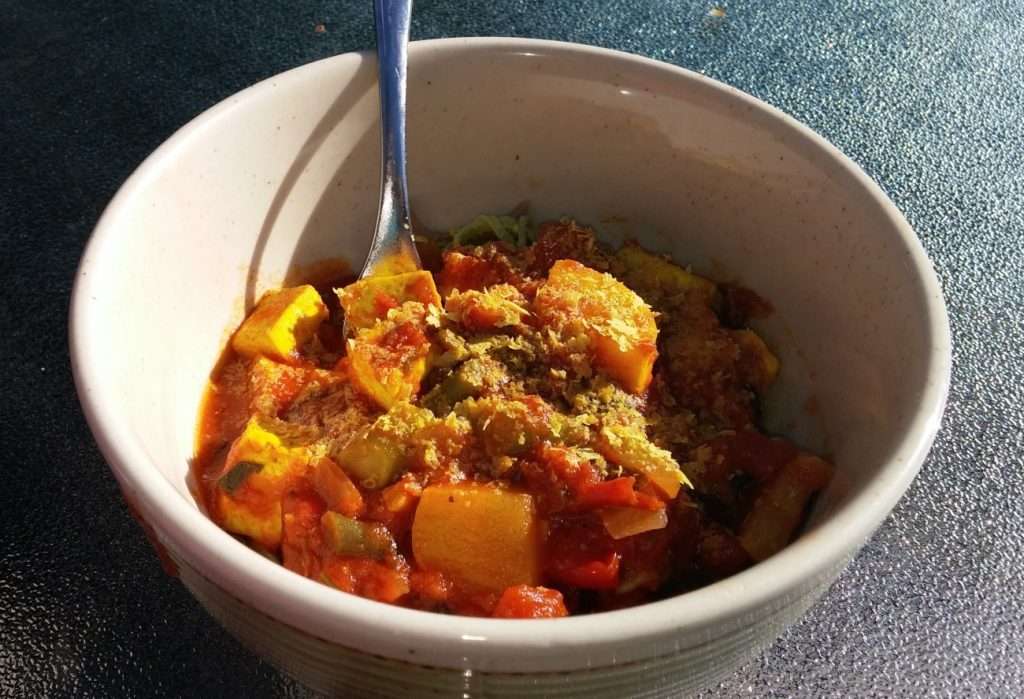 A bowl of plant-based vegetable stew with bright orange and yellow chunks, likely squash or peppers, topped with herbs. A spoon is placed inside the bowl. The vegan dish sits on a textured, dark surface, reflecting the careful guidance of a dietitians expertise.