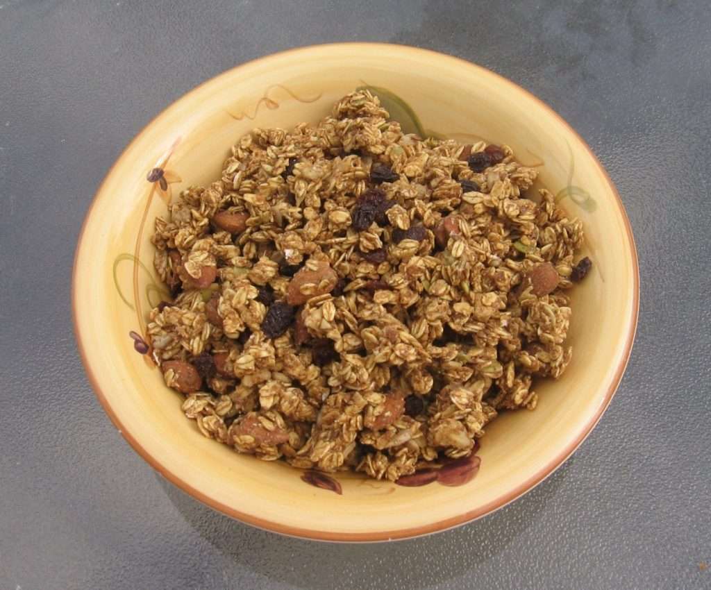 A bowl of vegan granola sits on a glass table. The chunky mix includes visible almonds and raisins throughout. The beige bowl features subtle decorative patterns on the rim, enhancing its plant-based appeal.