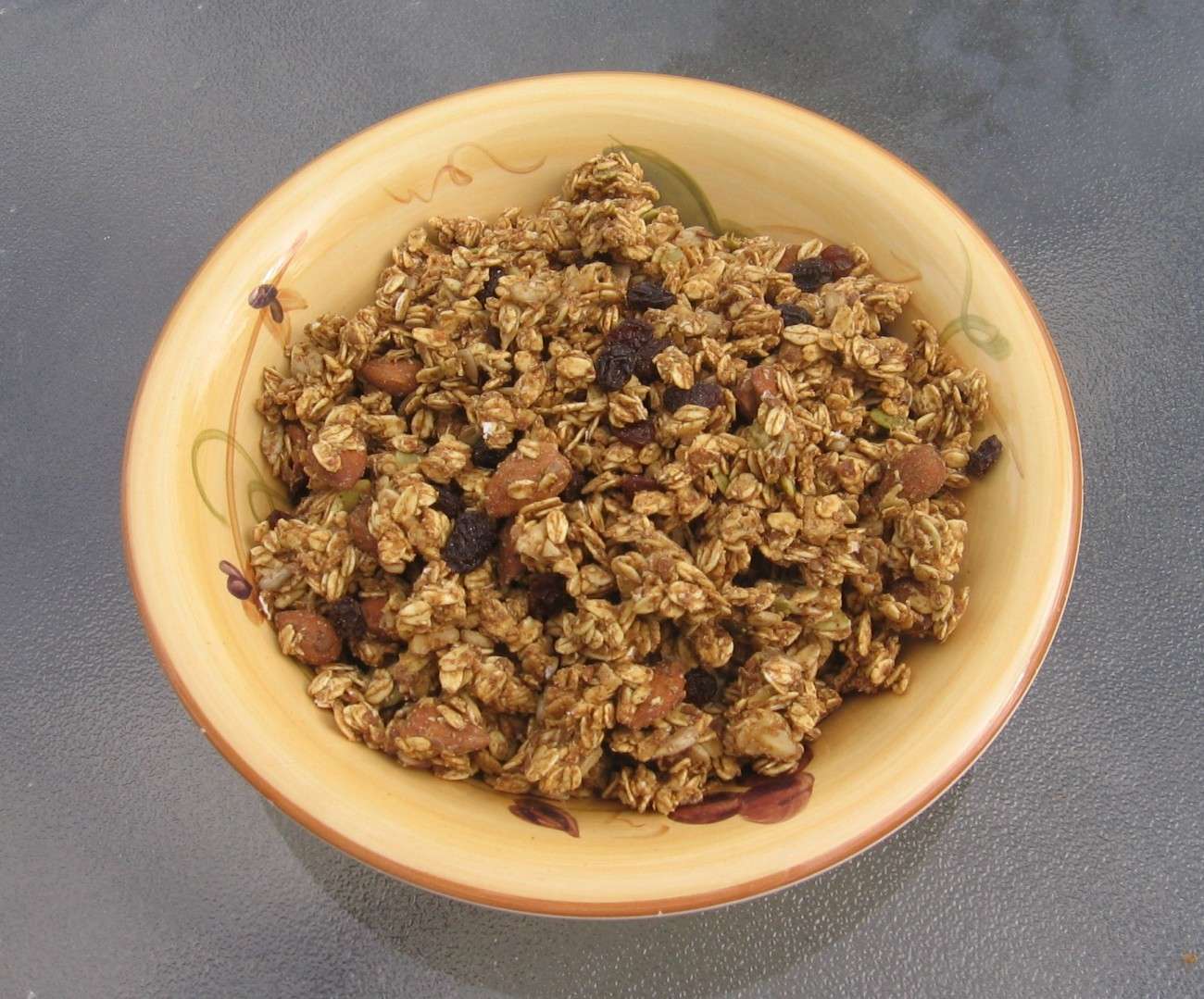 A bowl of vegan granola sits on a glass table. The chunky mix includes visible almonds and raisins throughout. The beige bowl features subtle decorative patterns on the rim, enhancing its plant-based appeal.