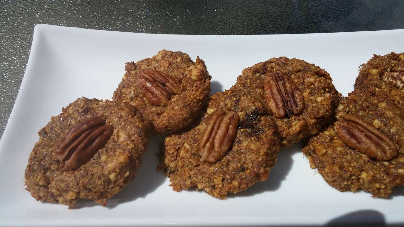 Five nutty, plant-based cookies with pecan halves on top are arranged on a rectangular white plate. The cookies appear to be homemade, with a rustic texture, and the plate is set on a textured surface. The lighting suggests its a sunny day.