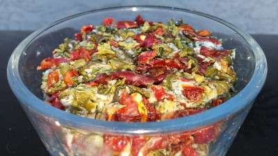 A clear glass bowl filled with a vibrant, plant-based mix of dried herbs and flowers, including red petals and green leaves, against a neutral background.