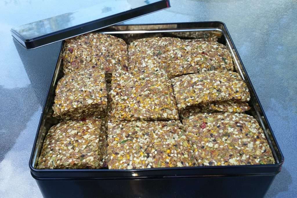 A black metal tin filled with square, seed-covered crackers showcases a delightful vegetarian treat. The lid is propped open on the side. Packed with various seeds and grains, these plant-based wonders boast a textured appearance as they sit on a reflective surface.
