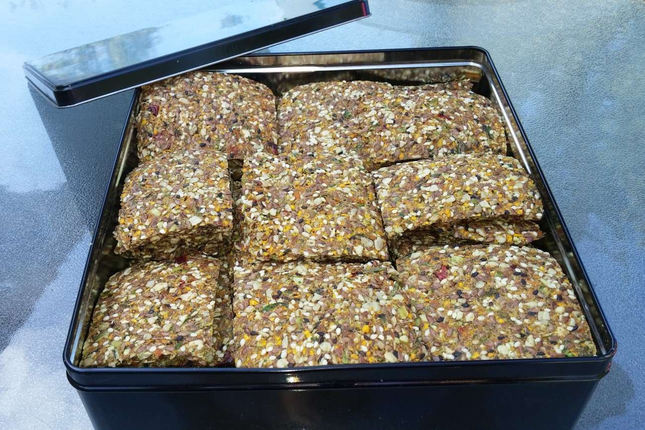 A black metal tin filled with square, seed-covered crackers showcases a delightful vegetarian treat. The lid is propped open on the side. Packed with various seeds and grains, these plant-based wonders boast a textured appearance as they sit on a reflective surface.