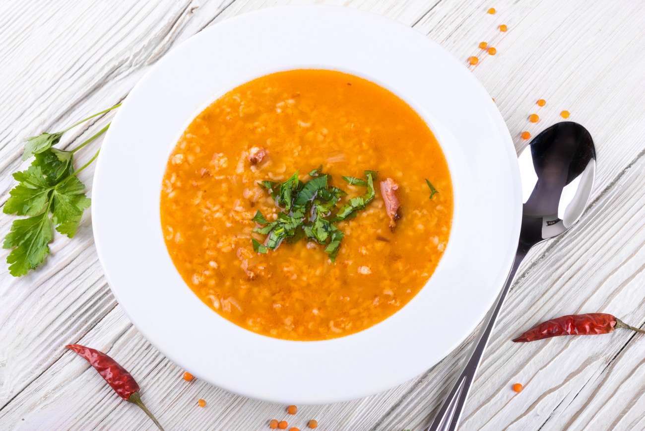 A bowl of plant-based lentil soup with chopped herbs on top, served in a white dish. A spoon, red chili peppers, and parsley are artfully placed around the bowl on a wooden surface.