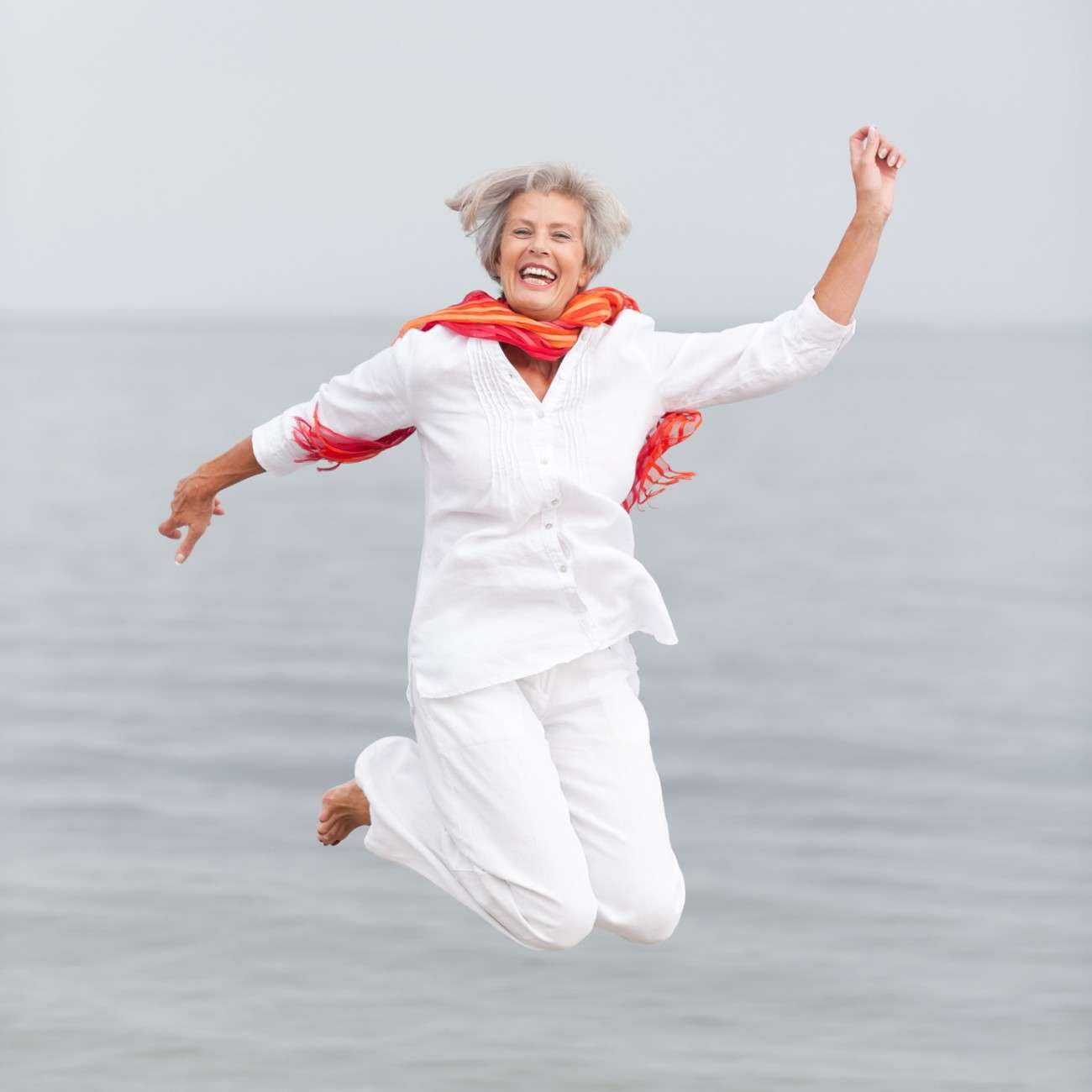 An elderly woman, embracing a vibrant vegetarian lifestyle, joyfully jumps in the air on a beach, wearing a white outfit and red scarf with the sea stretching behind her.