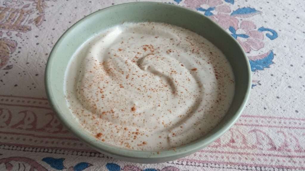 A green bowl filled with creamy yogurt, sprinkled with cinnamon, sits on a patterned tablecloth adorned with floral and decorative designs. This delightful treat is perfect for a vegetarian dietitian seeking a delicious, plant-based snack.
