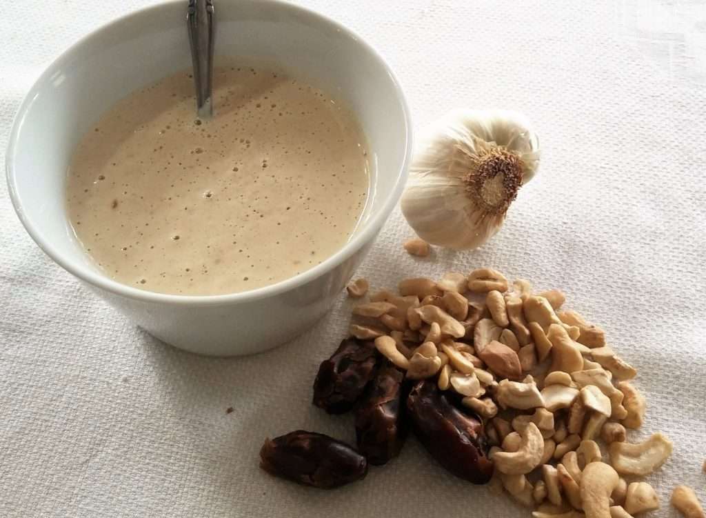 A bowl of creamy beige sauce, perfect for a vegetarian or vegan dish, sits with a spoon on a white surface. Nearby are a garlic bulb, cashews, and dates, showcasing wholesome ingredients that any dietitian would approve of.