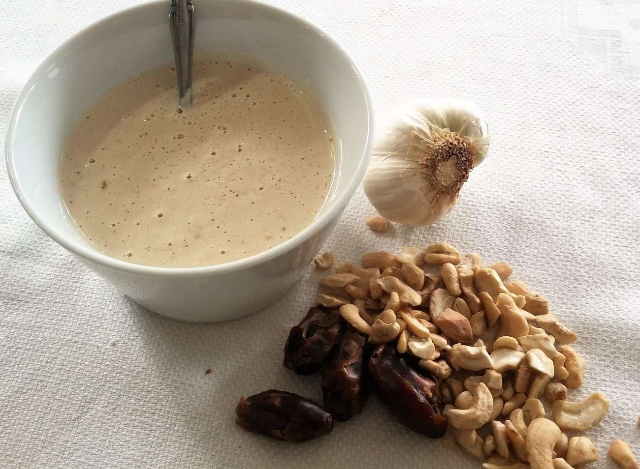 A bowl of creamy beige sauce, perfect for a vegetarian or vegan dish, sits with a spoon on a white surface. Nearby are a garlic bulb, cashews, and dates, showcasing wholesome ingredients that any dietitian would approve of.