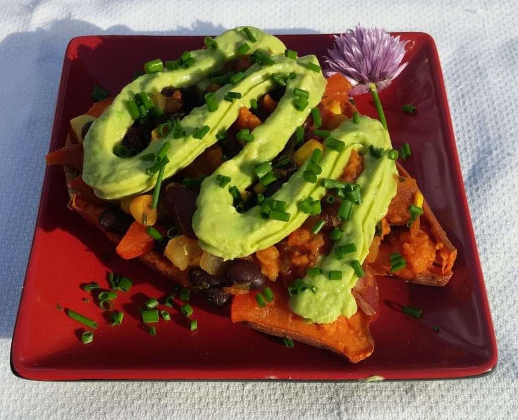 A red plate holds a delicious vegan baked sweet potato topped with black beans, corn, chopped vegetables, and a generous drizzle of creamy avocado sauce. Chopped chives and a purple edible flower garnish this vibrant plant-based dish.