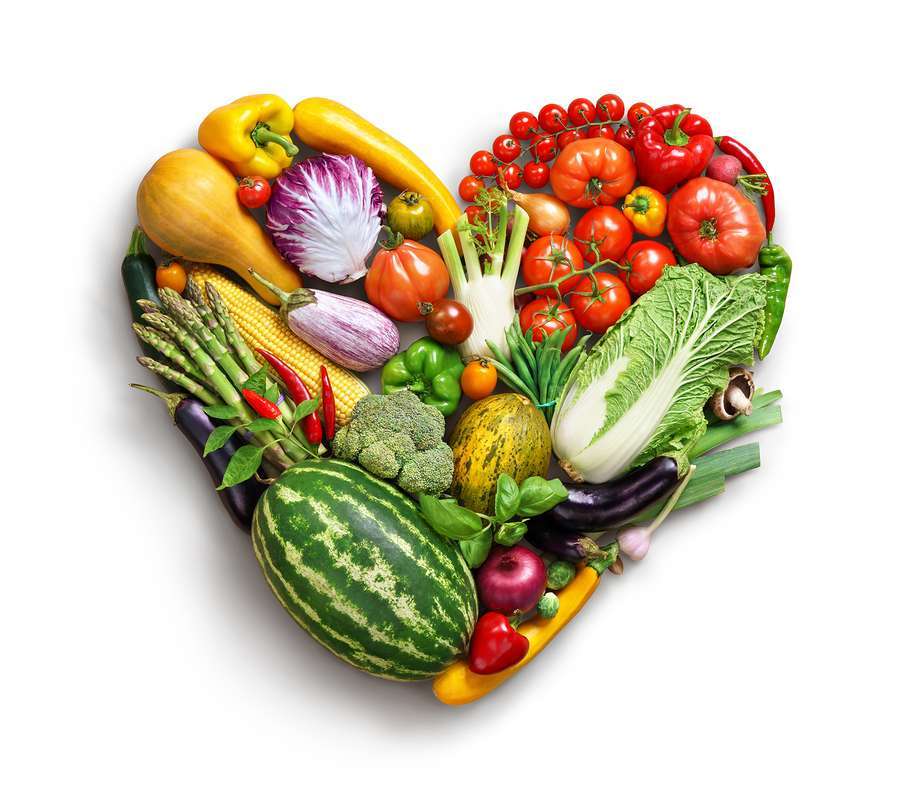 A heart-shaped arrangement of various vegetables and fruits, including tomatoes, watermelon, asparagus, carrots, broccoli, cabbage, corn, eggplant, bell peppers, and lettuce against a white background—perfect for any vegan or vegetarian dietitian promoting healthy eating habits.