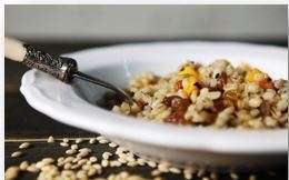 A white plate filled with a vibrant, plant-based grain salad, featuring ingredients like corn and beans, is set on a wooden surface. A fork rests on the edge of the plate while some grains are playfully scattered around.