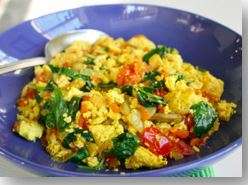 A colorful, vegan dish of scrambled tofu with spinach, tomatoes, and diced vegetables is served on a blue plate. A silver spoon rests on the side.