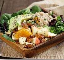 A wooden bowl filled with a vibrant, plant-based salad featuring fresh spinach, sliced avocado, quinoa, apple chunks, orange segments, and almonds. This vegetarian delight is sprinkled with seeds and elegantly set on a woven placemat.