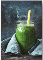 A plant-based green smoothie in a mason jar with a yellow straw sits on a dark surface, partially wrapped in a blue cloth. The textured background enhances its fresh and vibrant appeal.