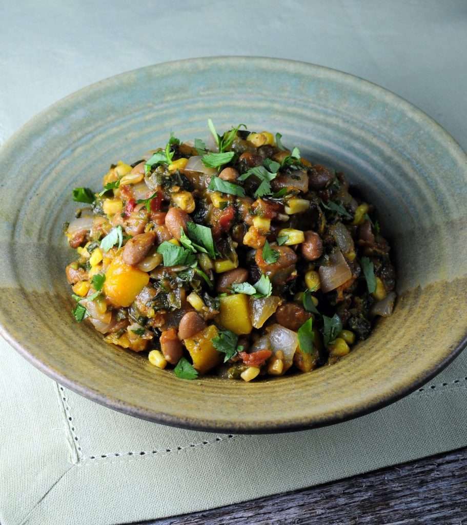 A ceramic bowl brimming with a vibrant, plant-based vegetable stew featuring beans, corn, diced squash, onions, and leafy greens. Garnished with chopped herbs, it sits elegantly on a light green tablecloth.