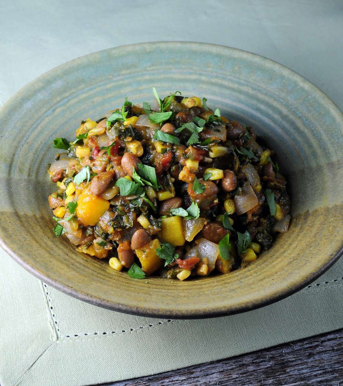A ceramic bowl brimming with a vibrant, plant-based vegetable stew featuring beans, corn, diced squash, onions, and leafy greens. Garnished with chopped herbs, it sits elegantly on a light green tablecloth.
