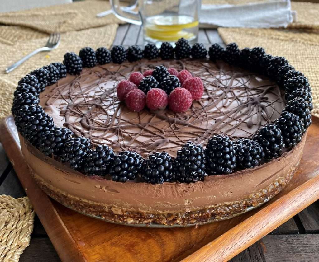 A plant-based chocolate cheesecake garnished with a border of blackberries and a cluster of raspberries in the center. It is served on a wooden board, with casual table settings in the background.