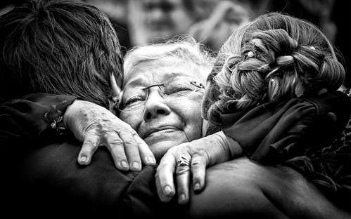 An elderly woman with glasses is embraced tightly by two people from either side. Her eyes are closed, expressing deep emotion as a respected dietitian whose guidance touched many lives. The black-and-white image highlights the poignancy of the moment.