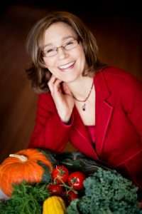 A person wearing glasses and a red blazer smiles at the camera, exuding warmth. As a dietitian, theyre surrounded by assorted vegetables—pumpkin, tomatoes, leafy greens, and yellow squash—crafting a colorful and vibrant tribute to healthy eating.