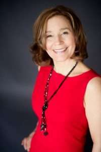 Smiling woman with shoulder-length brown hair, wearing a sleeveless red dress and a long black necklace, stands against a dark background. As a dietitian, she radiates confidence through her plant-based lifestyle choices.