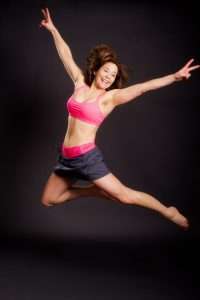 A woman wearing a pink sports bra and black shorts leaps joyfully against a dark background. Her arms and legs are extended, radiating energy like a plant-based enthusiast. Shes smiling vibrantly, one hand flashing a peace sign reminiscent of harmony and balance.