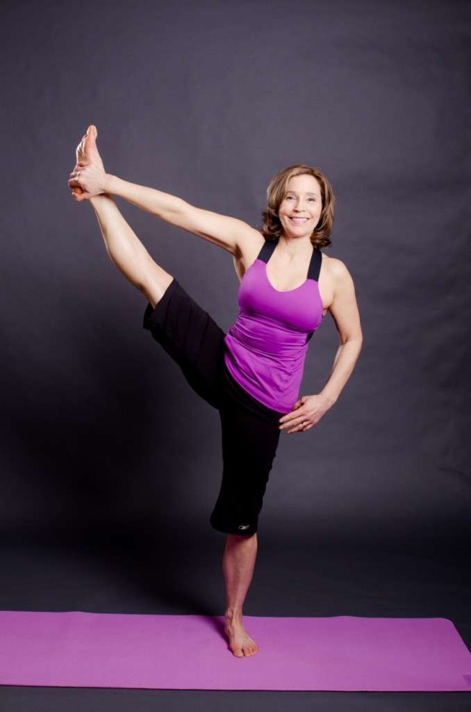 A woman in a purple tank top and black pants, embodying a vegan lifestyle, performs a standing yoga pose on a purple mat. She balances gracefully on one leg while extending the other sideways, holding her foot with her hand. The background is a plain dark gray.