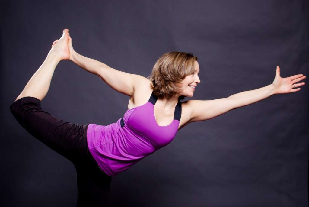 A person in a purple tank top and black pants gracefully balances in a yoga pose, embodying the serenity often associated with a plant-based lifestyle. Standing on one leg with the other extended back and an arm reaching forward, they exude harmony against the dark background.