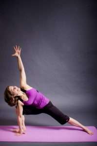 A person dressed in a purple tank top and black pants performs a side angle yoga pose on a pink mat, embodying the balance of a plant-based lifestyle. One arm extends upward while the other rests on the floor against the gray background, showcasing harmony and strength.