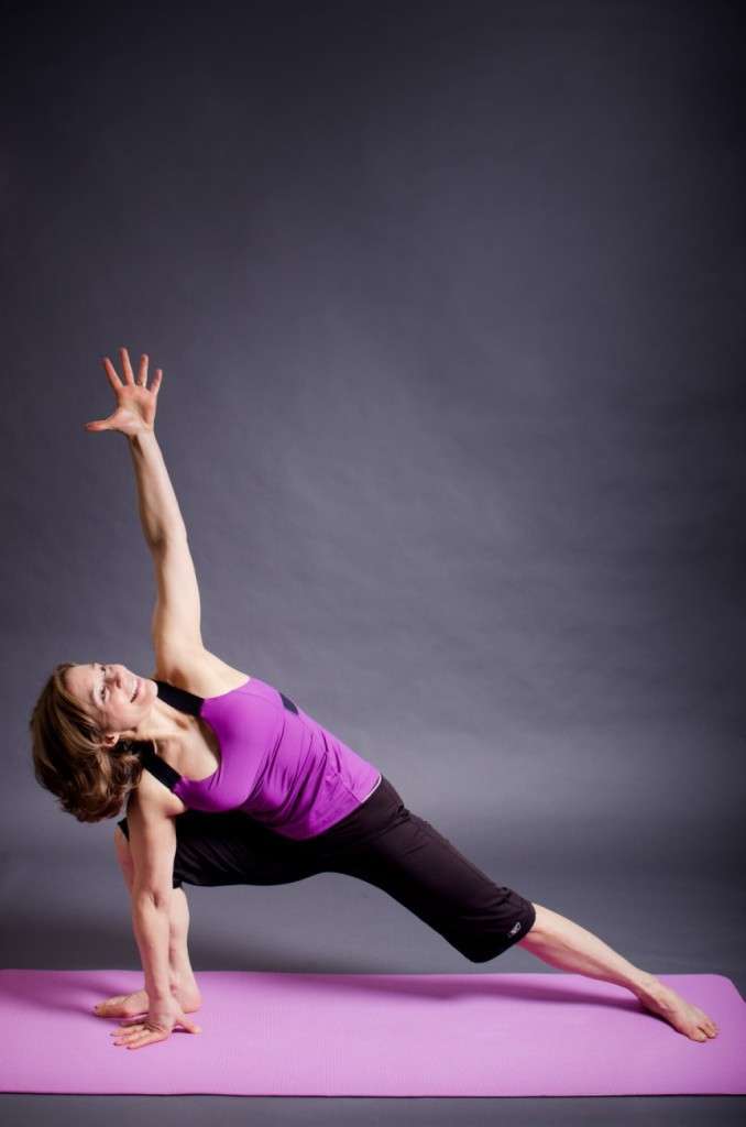 A person dressed in a purple tank top and black pants performs a side angle yoga pose on a pink mat, embodying the balance of a plant-based lifestyle. One arm extends upward while the other rests on the floor against the gray background, showcasing harmony and strength.