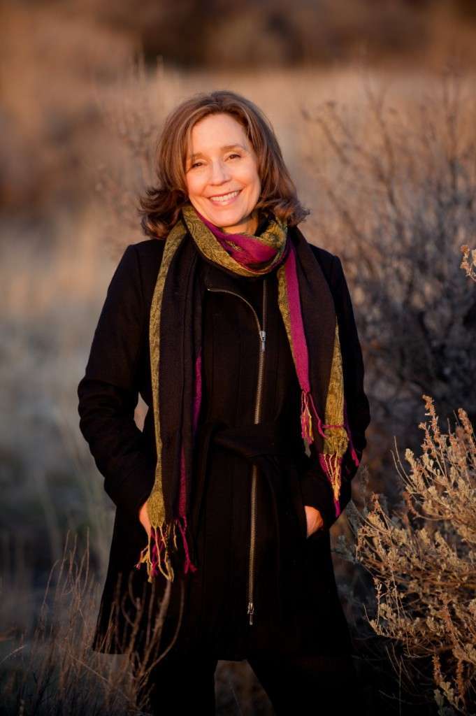 A woman stands outdoors, smiling with her hands in the pockets of a black coat. She sports a colorful scarf showcasing green and purple shades. The background—a blurred natural landscape with dry grasses and bushes—reflects the serene beauty of her plant-based lifestyle.