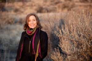 A woman with wavy brown hair smiles while standing outdoors in a natural, dry landscape. A dietitian by profession, she wears a black coat and a colorful scarf. The background includes shrubs and grasses bathed in warm, golden light.