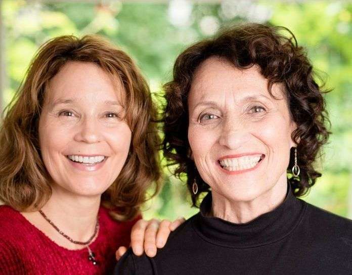 Two women, radiating health and joy, smile at the camera. The one with curly brown hair in a black top and earrings stands next to her friend in a red top with wavy hair and a necklace. Both share a passion for a plant-based lifestyle, set against a blurred, green backdrop.