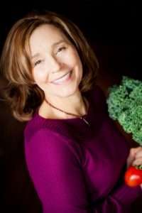 A smiling woman in a purple sweater, embodying the essence of a plant-based lifestyle, holds a bunch of kale and a red tomato, set against a dark background.