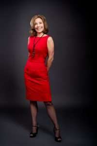 A woman stands confidently against a dark background, wearing a sleeveless red dress and black heels. As a dietitian, she radiates positivity with her shoulder-length hair and warm smile, while her stance reflects the vibrant energy of a plant-based lifestyle.