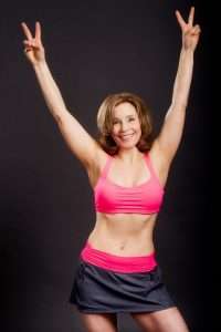 A person wearing a bright pink sports bra and dark shorts stands against a dark background with arms raised in a V shape, smiling energetically, embodying the vibrant spirit of a dedicated vegan.